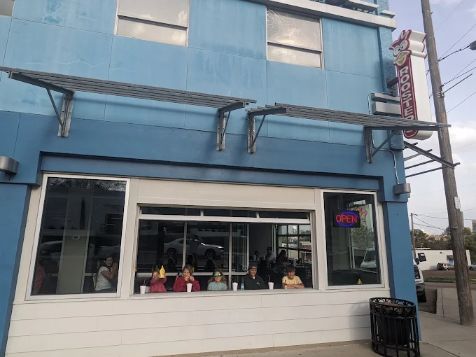 A closer look at Rooster’s open-air facade, showing the vintage neon sign, people walking by, and the large garage-style windows creating a neighborly atmosphere.